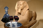בקבוק יין מפואר עם ארבע כוסות יין על מגש, ברקע פסל מצרי עתיק